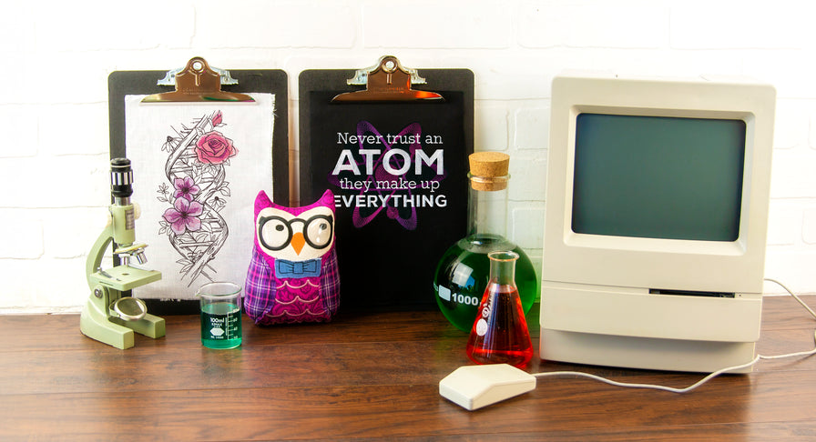 Back to School embroidery display featuring a vintage computer, microscope, and science-themed props. Two embroidered clipboards show designs from the Ghost Baroque collection, including a floral skeleton and the phrase “Never trust an atom, they make up everything.” A pink owl plushie with glasses sits in front.
