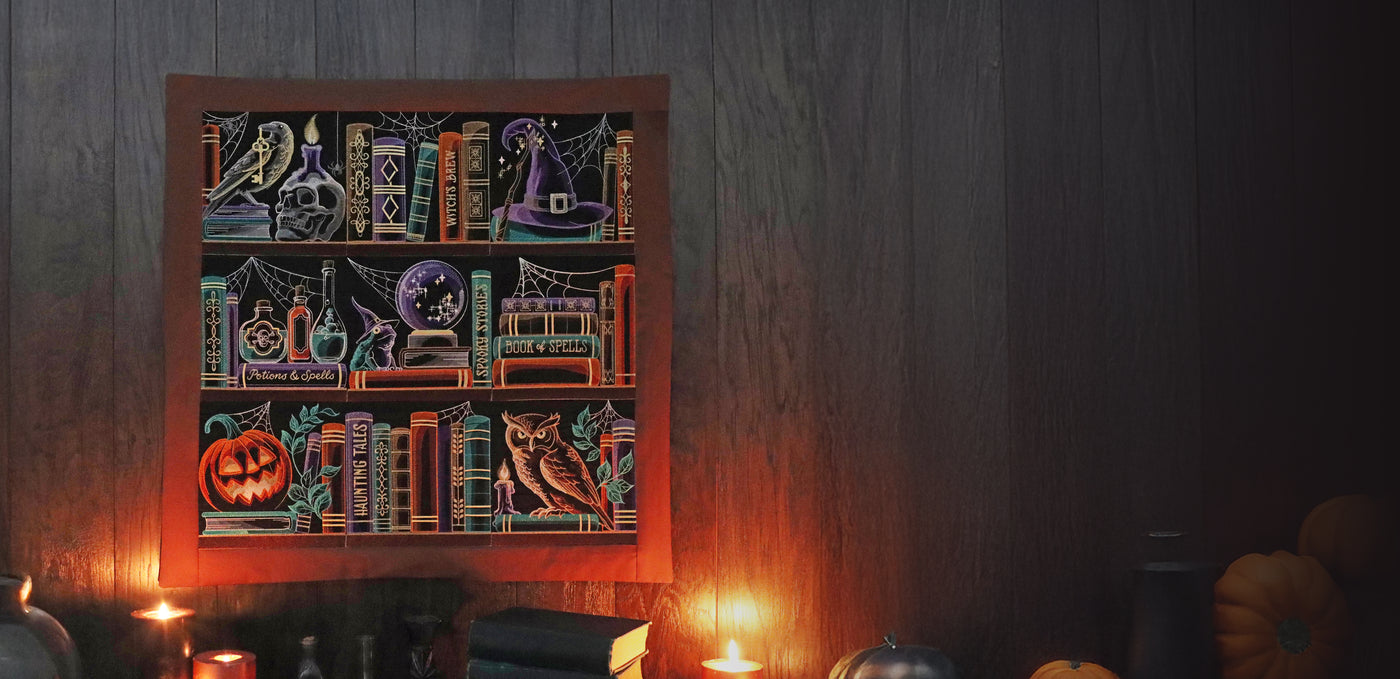 Halloween embroidery wall hanging with books, potions, pumpkin, owl, and witch hat. Surrounded by candles, pumpkins and books.