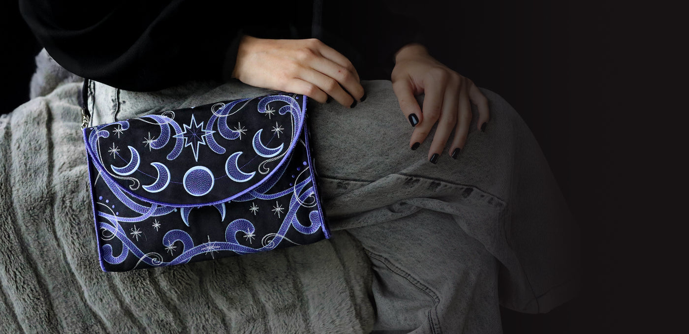 Person holding a black machine embroidered clutch with celestial designs on a dark background
