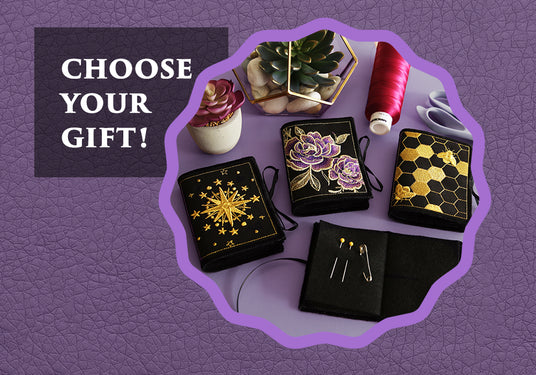 hree black felt needle books embroidered with gold starburst, purple roses, and honeycomb with bee designs, displayed with thread, scissors, and succulents, next to text that reads “Choose Your Gift!”
