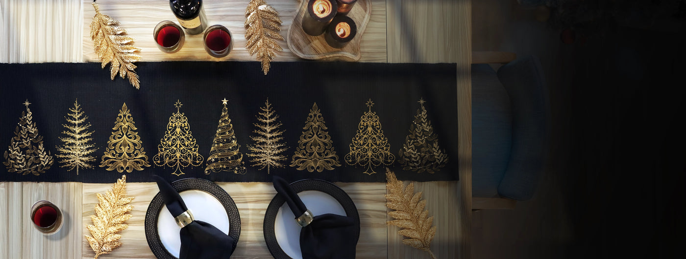 Decorative machine embroidery table runner with gold Christmas tree designs on a black background, accompanied by wine glasses and bottles.