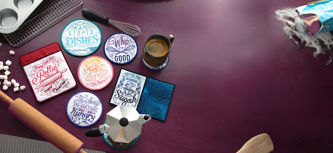 Set of colorful machine embroidery in-the-hoop kitchen set with humorous sayings on a dark surface with kitchen items.