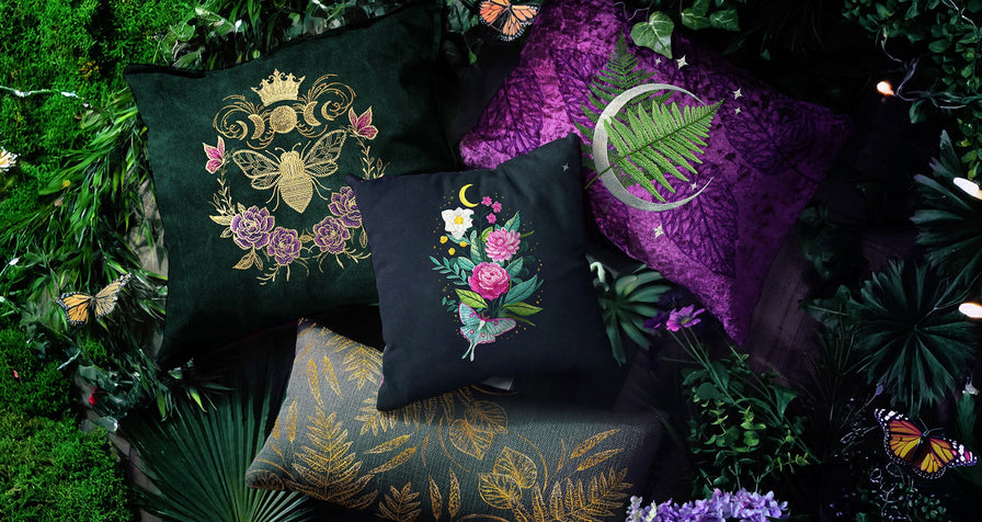 Decorative pillows with nature-inspired machine embroidery designs, including floral motifs, bees, ferns, and a moth under a crescent moon, displayed against a lush garden backdrop with butterflies and greenery.