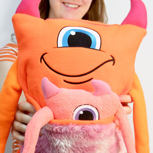 Smiling woman holds two plush toys featuring monster-themed embroidery—one orange with horns and one small pink creature.