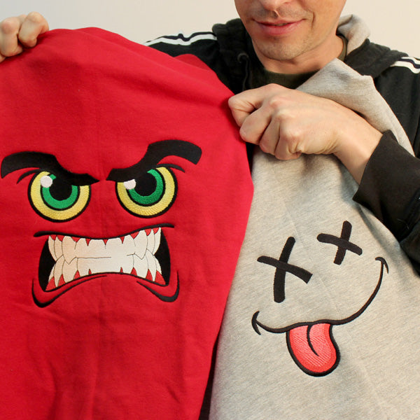 Two shirts display embroidered Monster Factory faces: one angry, one with its tongue out; both show stitching detail.
