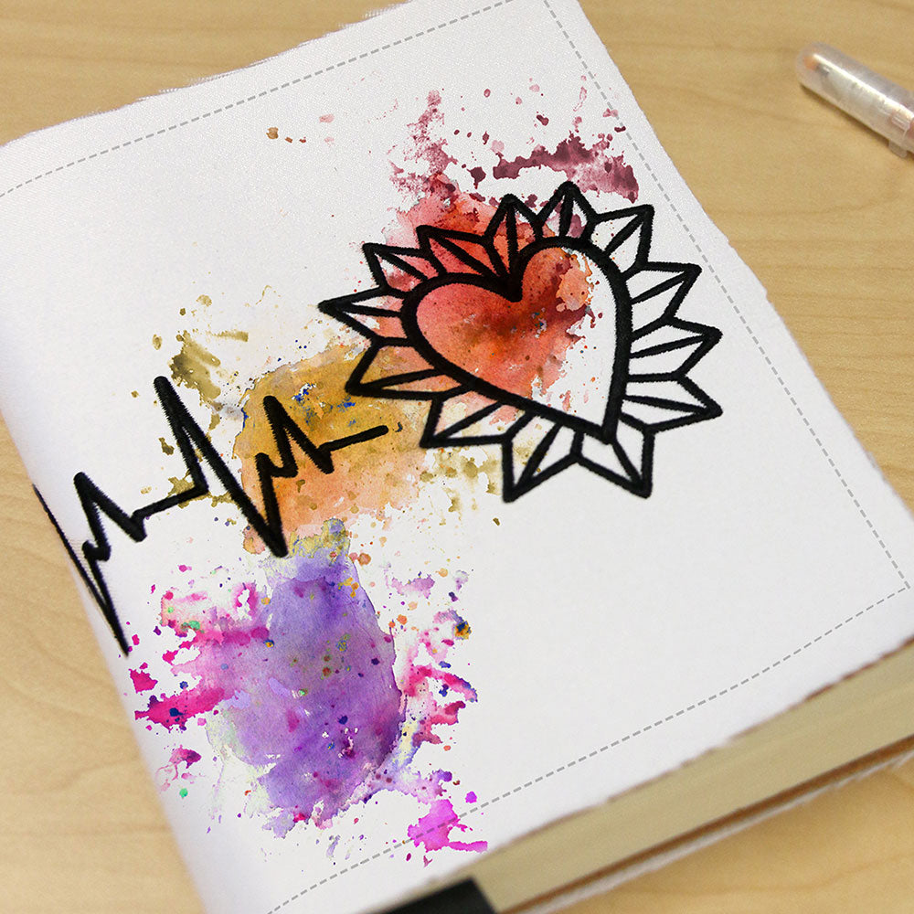 Embroidery design features colorful splatters, a black heart, sunburst, and heartbeat line on notebook covers.