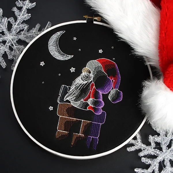 Embroidery hoop with stitched image of Santa Claus in a red hat sitting on a brown chimney under a crescent moon and stars on black fabric.