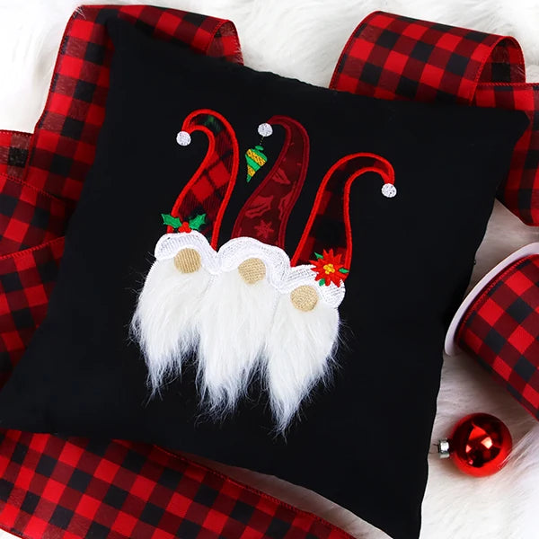 Black pillow with three embroidered gnomes wearing red and patterned hats, white beards, and beige noses, surrounded by red and black plaid ribbon and a red ornament.