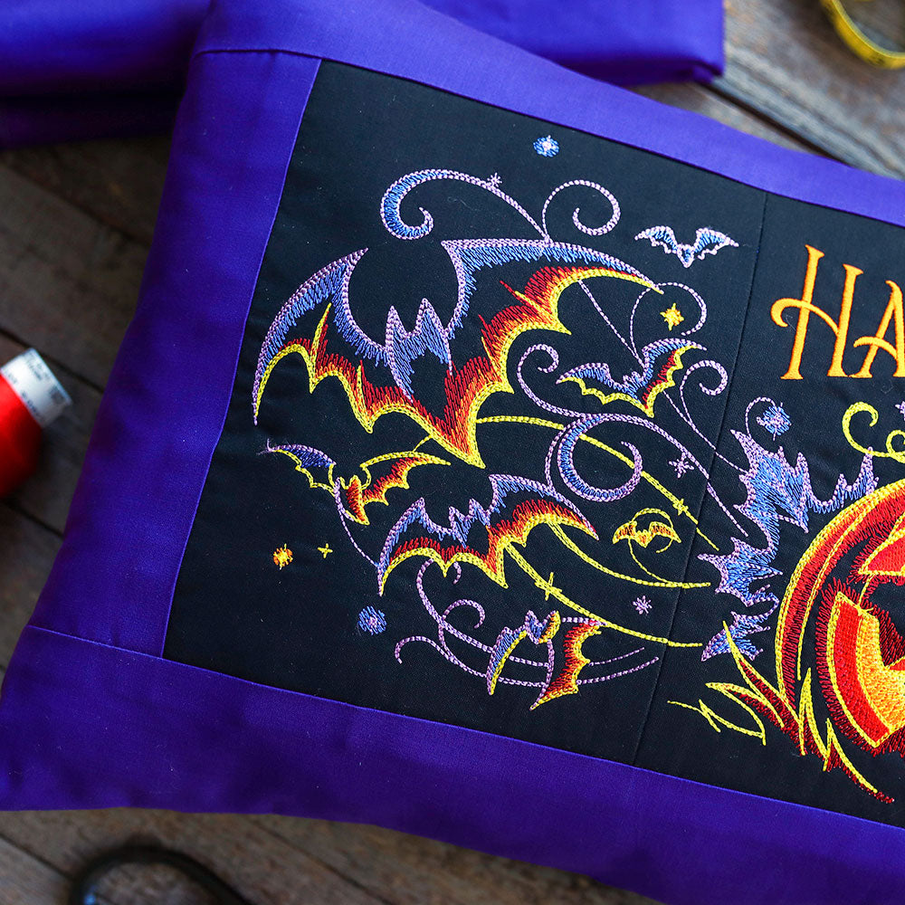 Black pillow featuring embroidered bats, stars, pumpkins, and the words Happy Halloween in festive lettering.