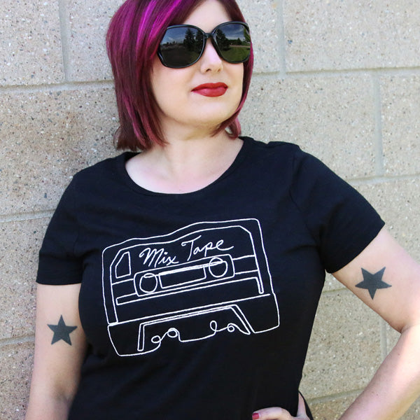 Person wearing a black t-shirt with a machine embroidered cassette tape and 'Mix Tape' text design, standing against a brick wall.