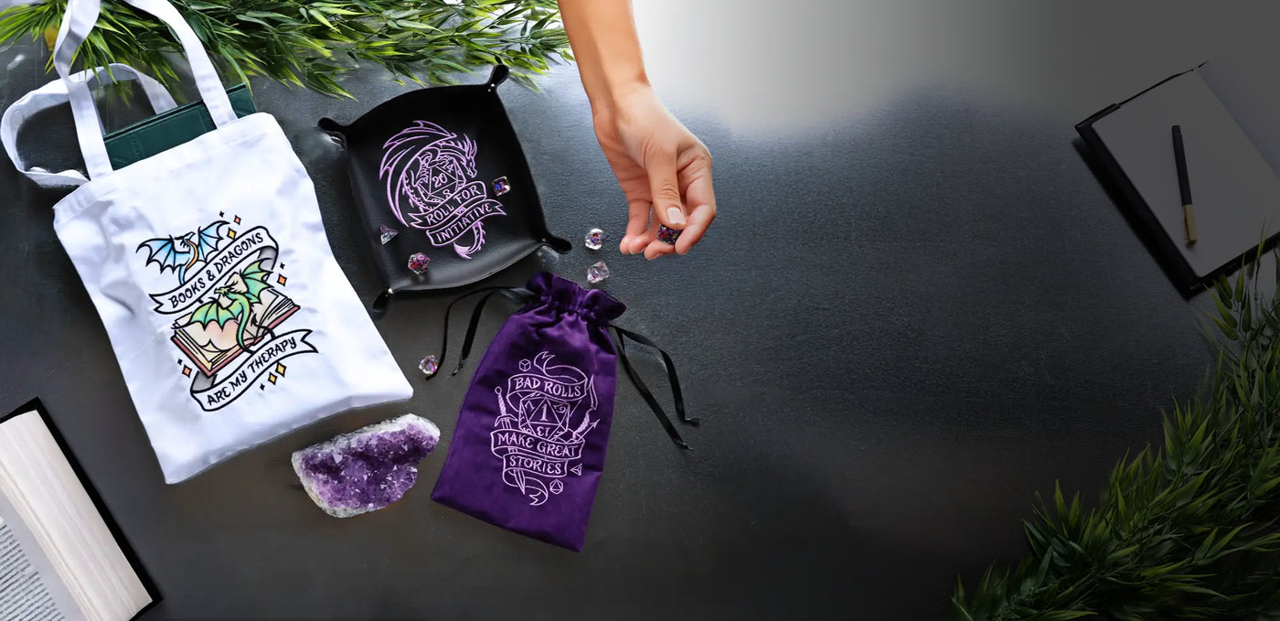 Machine embroidered, dice bag, dice tray and tote bag with machine embroidery designs staged on a dark surface with a hand holding dice.