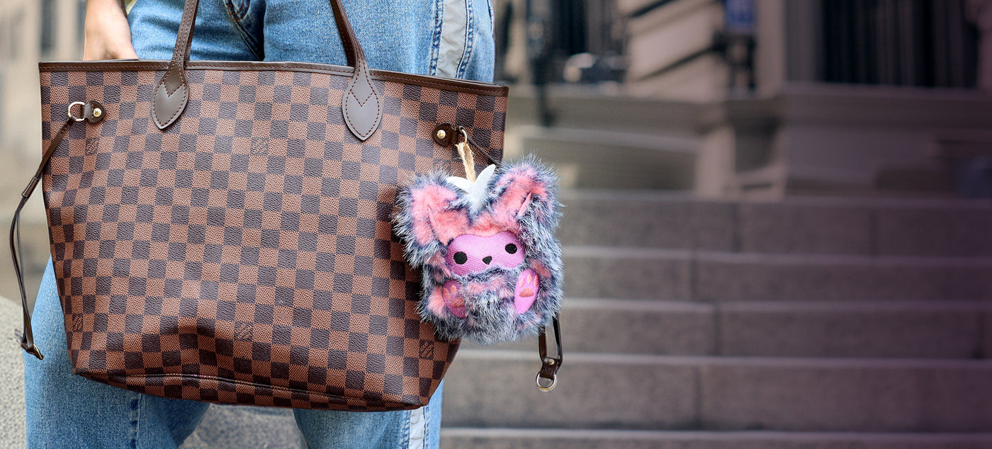 Checkered handbag with a plush machine embroidery monster keychain, held by a person wearing jeans, on a blurred background.