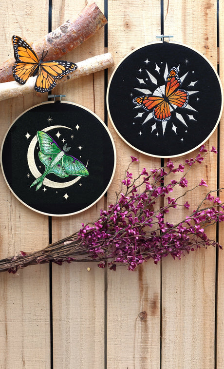 Two embroidery hoops with machine embroidery monarch butterfly and luna moth designs on a wooden surface with purple flowers.