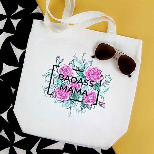 White tote bag with 'BADASS MAMA' machine embroidery design and sunglasses on a yellow and black patterned surface