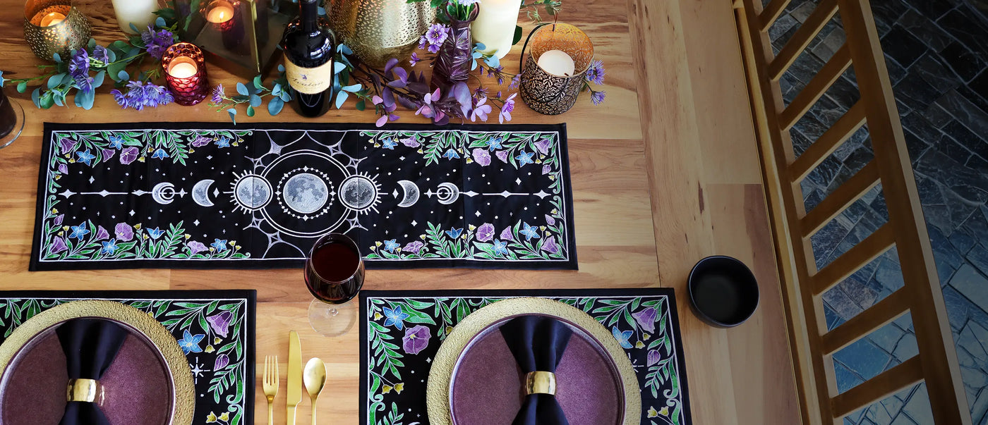 Decorative table setting with a machine embroidered celestial & floral themed multi-piece table runner & placemats, purple napkins, and candles on a wooden table.