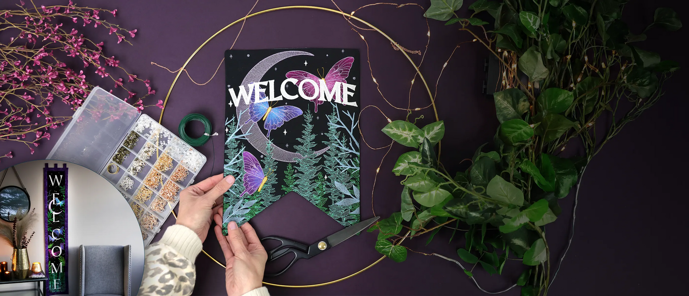 Decorative machine embroidered flag with the word 'Welcome' and nature-themed cover on a dark surface with plants and decorative items.