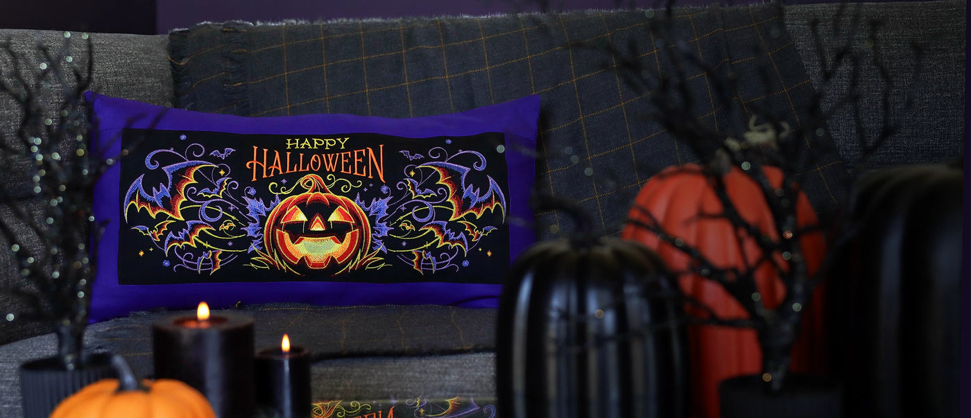 Halloween-themed pillow with jack-o'-lantern machine embroidery design on a couch with pumpkins and candles.
