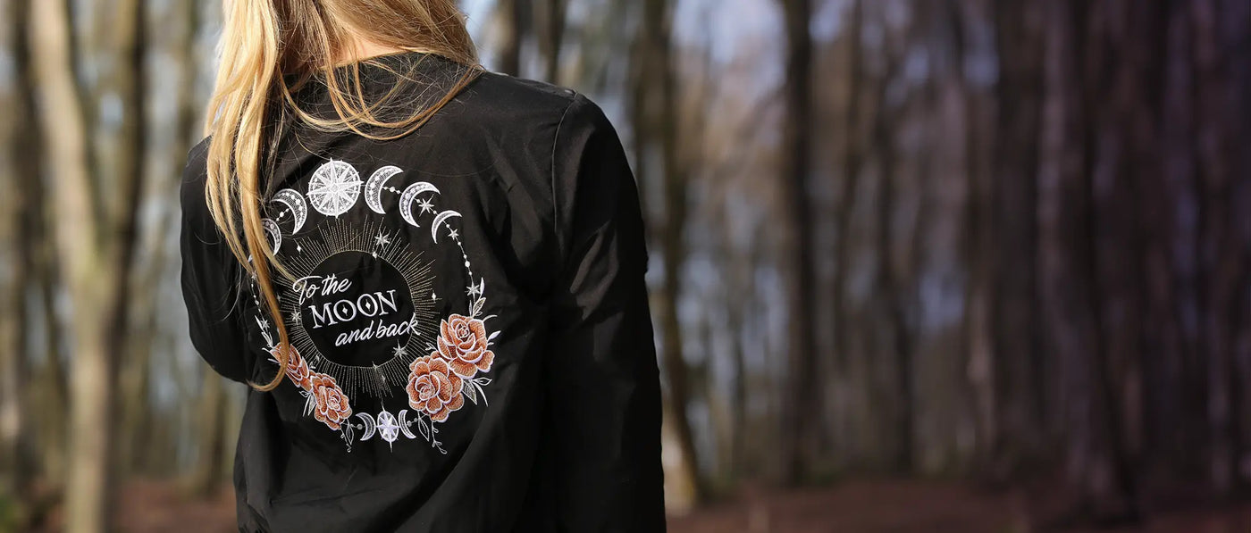 Person wearing a black jacker with machine embroidered moon phases and floral design stitched on the back in a forest setting.