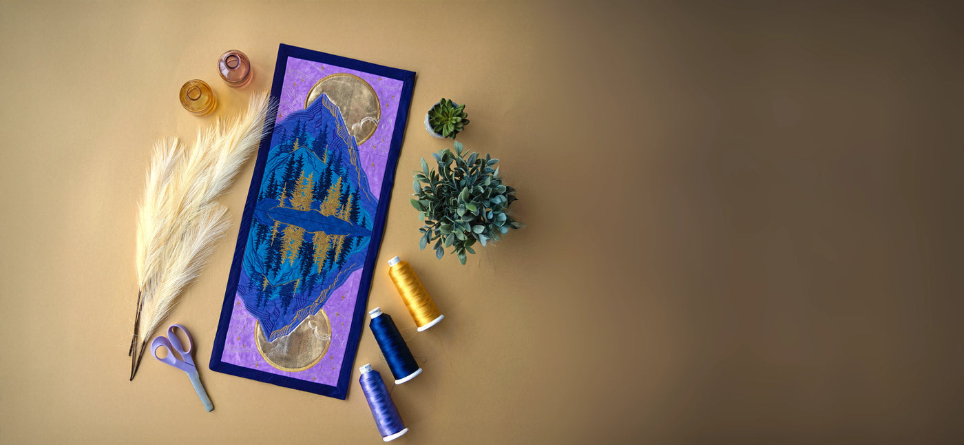 Machine embroidered mountain panel scene surrounded by thread, greenery and feathers.