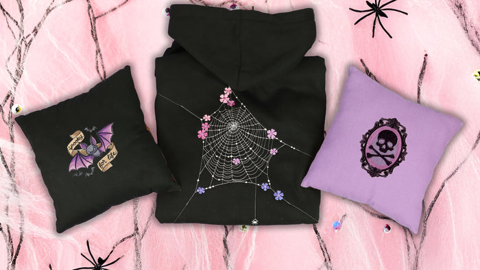 Black hoodie with machine embroidered floral spiderweb and black and purple pillows with a machine embroidered bat and skull designs on a pink background.