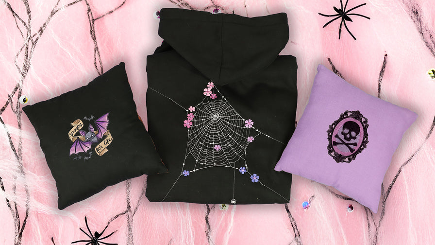 Black hoodie with spider web design, black pillow with purple bat design, and purple pillow with skull design on a pink background.