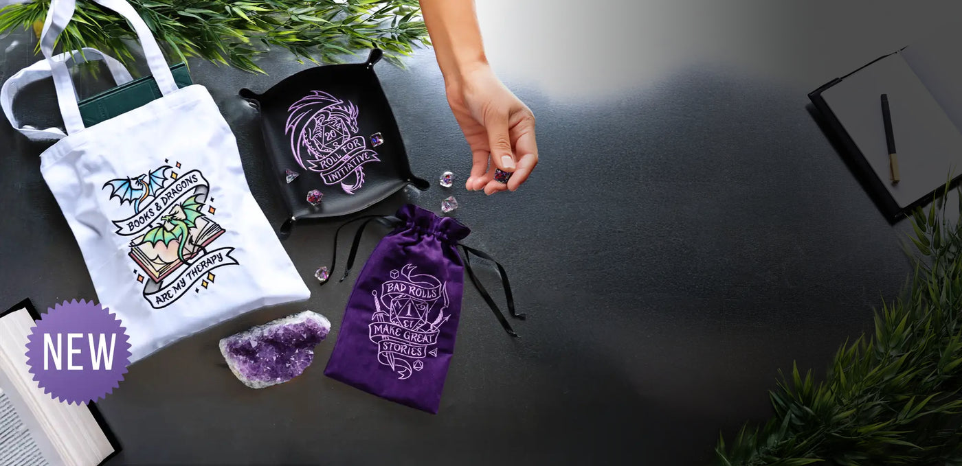 White tote bag with 'Books & Dragons are my Therapy' design, purple dice tray with dragon emblem, purple drawstring bag with text, and amethyst crystal on a dark surface.