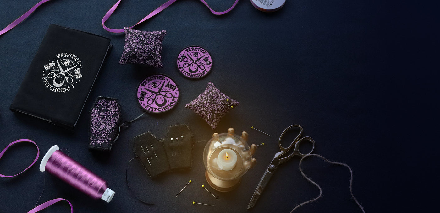 Embroidered pin cushion, needle book, patches and journal with "practice stitchcraft" text surrounded by a small lamp on a dark surface