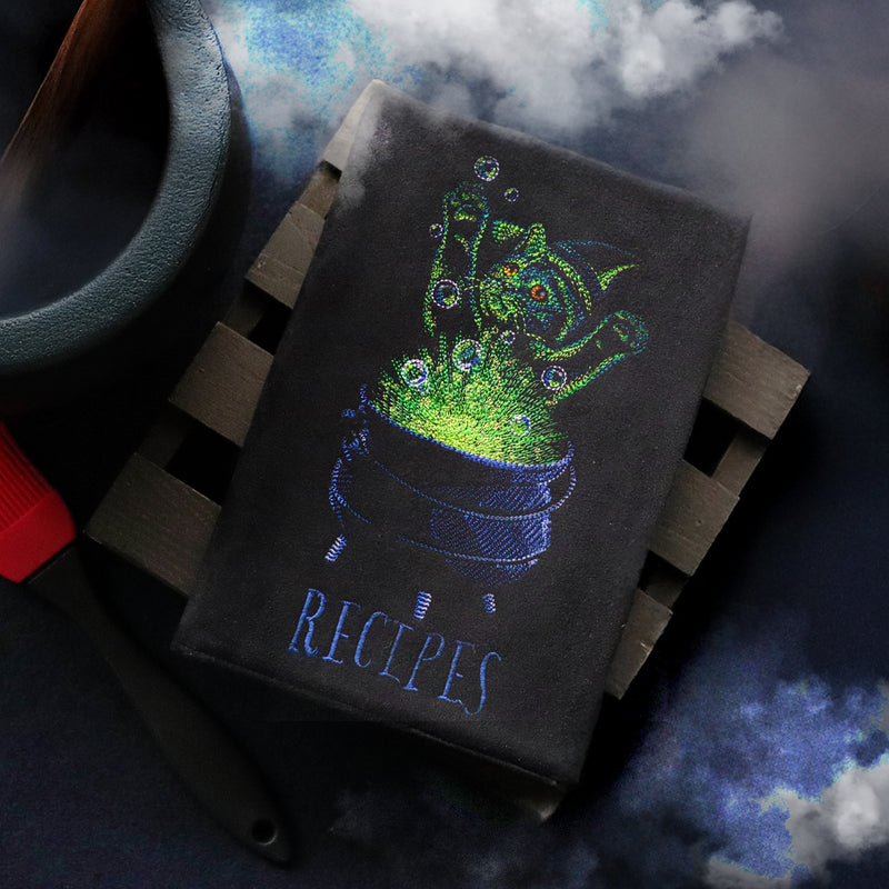 Black embroidered book with a green glowing cauldron design and 'RECIPES' text, on a dark background.