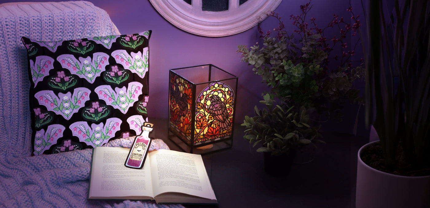 Decorative pillow with a bat pattern, stained glass raven design on lamp, and open book on a purple blanket.