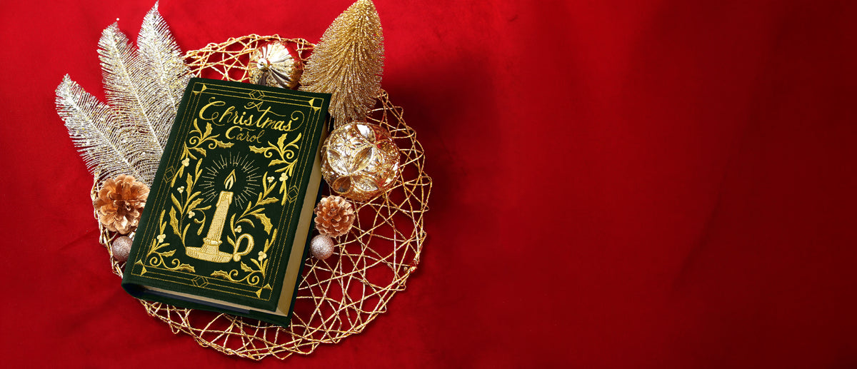 A green book with gold embossed lettering "A Christmas Carol" and candle design lies on a gold wire placemat surrounded by gold pinecones and decorative feathers.