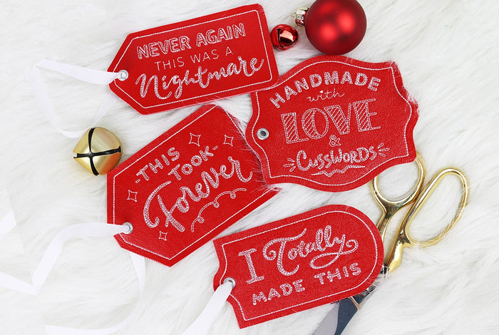 Machine embroidered red gift tags with humorous text on a white background with Christmas decorations.