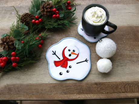 Machine embroidered Snowman coaster with a mug of hot chocolate on a wooden surface with Christmas decorations.