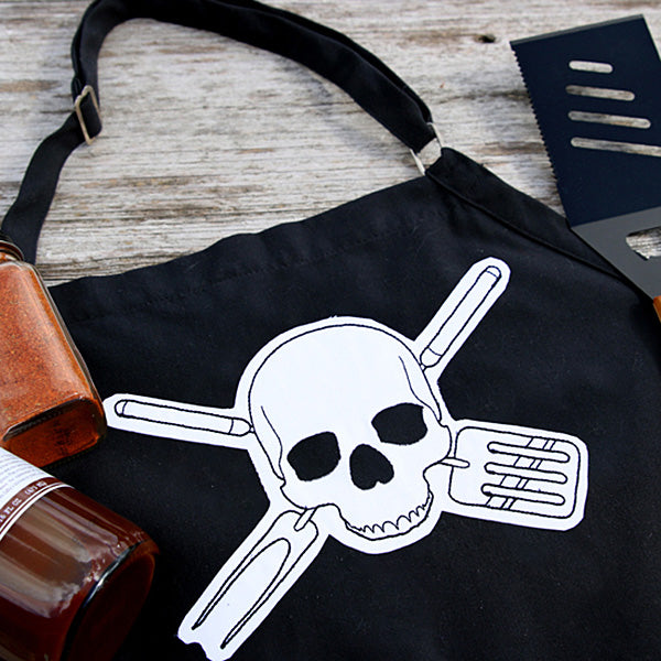 Black apron with applique machine embroidered skull and crossed spatulas design on a wooden surface with a bottle and spatula.
