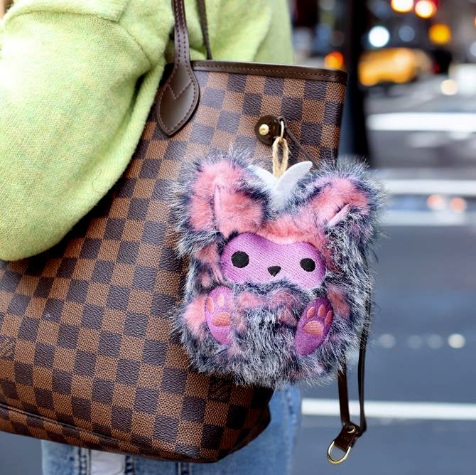 Checkered handbag with a fluffy pink and gray machine embroidered monster keychain attached, blurred city street background