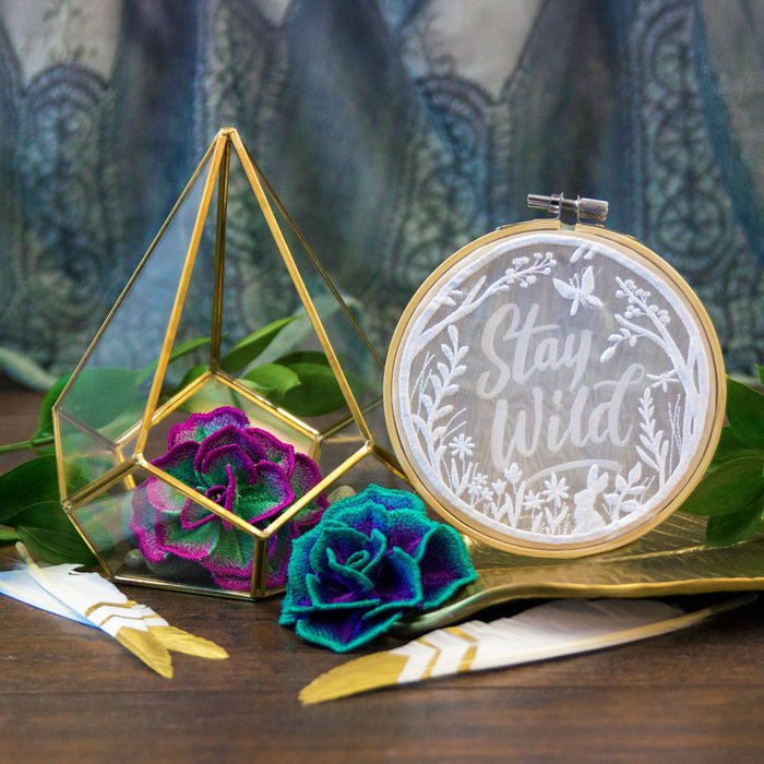Glass terrarium with colorful machine embroidered succulents  and embroidery hoop with 'Stay Wild' design on a wooden surface