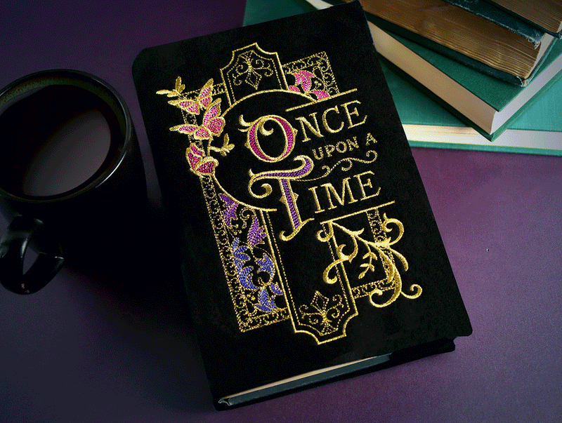 Black book with ornate machine embroidered “Once Upon a Time” cover design in gold, pink, and purple thread, styled beside stacked books and a coffee mug on a dark purple surface.