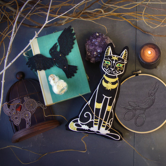Decorative machine embroidered items including a plush black cat with gold accents, freestanding lace crow and bunny, and a bird design in a hand embroidery hoop.