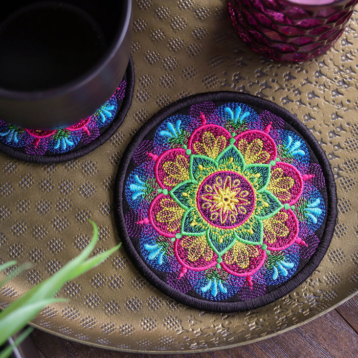 A round machine embroidered coaster with a vibrant multicolor mandala pattern sits on a textured gold tray next to a black cup, with another coaster partially visible in the background.