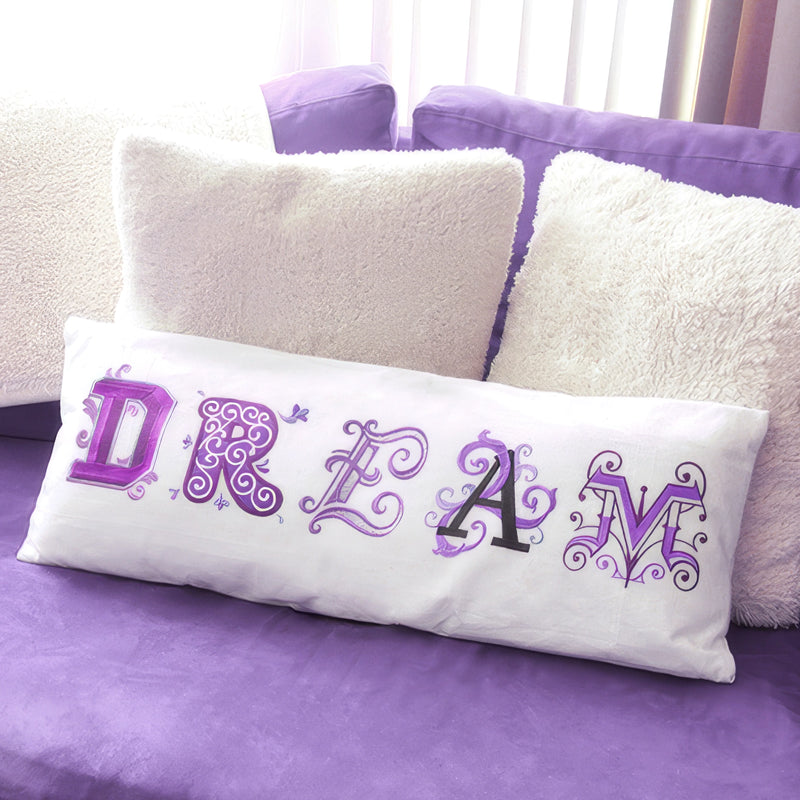 A white rectangular lumbar pillow with the word 'DREAM' spelled out in decorative machine embroidered purple and black letters, resting on a purple couch between fluffy cream pillows.