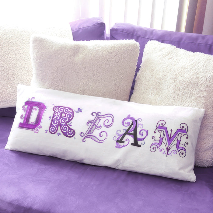 A white rectangular lumbar pillow with the word 'DREAM' spelled out in decorative machine embroidered purple and black letters, resting on a purple couch between fluffy cream pillows.