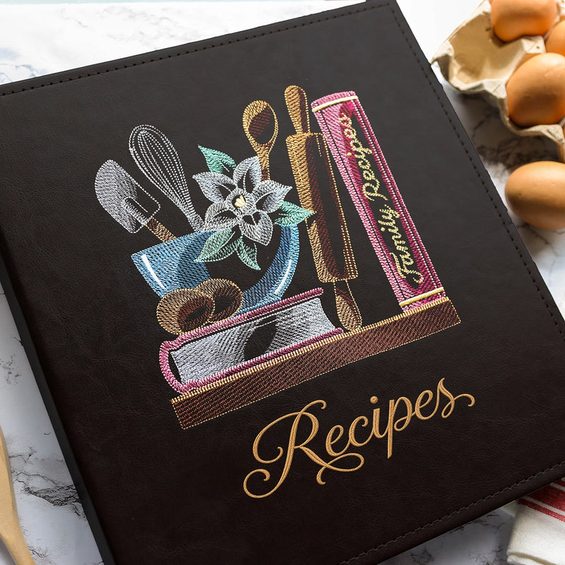 A black faux-leather recipe book with gold-embroidered 'Recipes' script on the cover, decorated with colorful embroidered kitchen utensils, a flower, and a pink cookbook. The book is styled on a marble surface with eggs nearby.