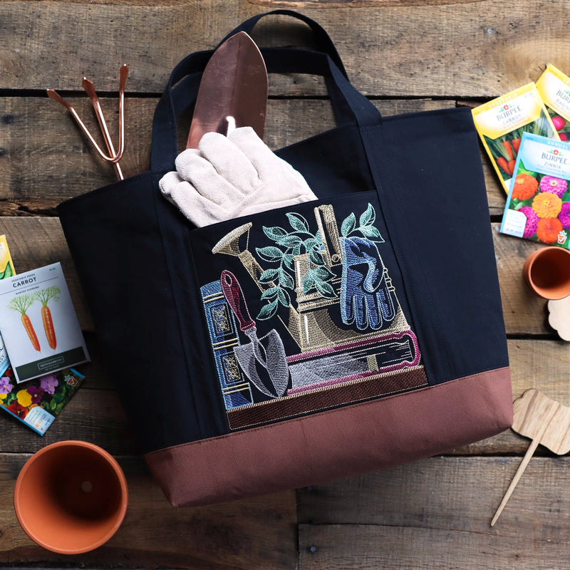 Black tote bag with gardening-themed machine embroidery design on a wooden surface with gardening tools and seeds.