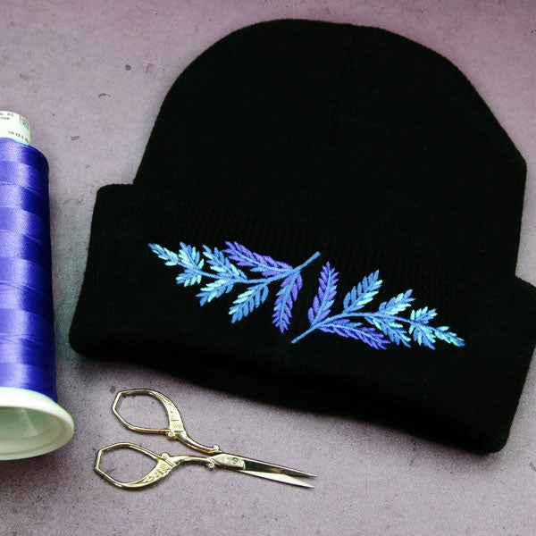 Black beanie with blue machine embroidered leaf design next to spool of thread and scissors on a gray surface