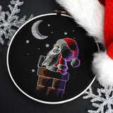 Embroidery hoop with stitched image of Santa Claus in a red hat sitting on a brown chimney under a crescent moon and stars on black fabric.