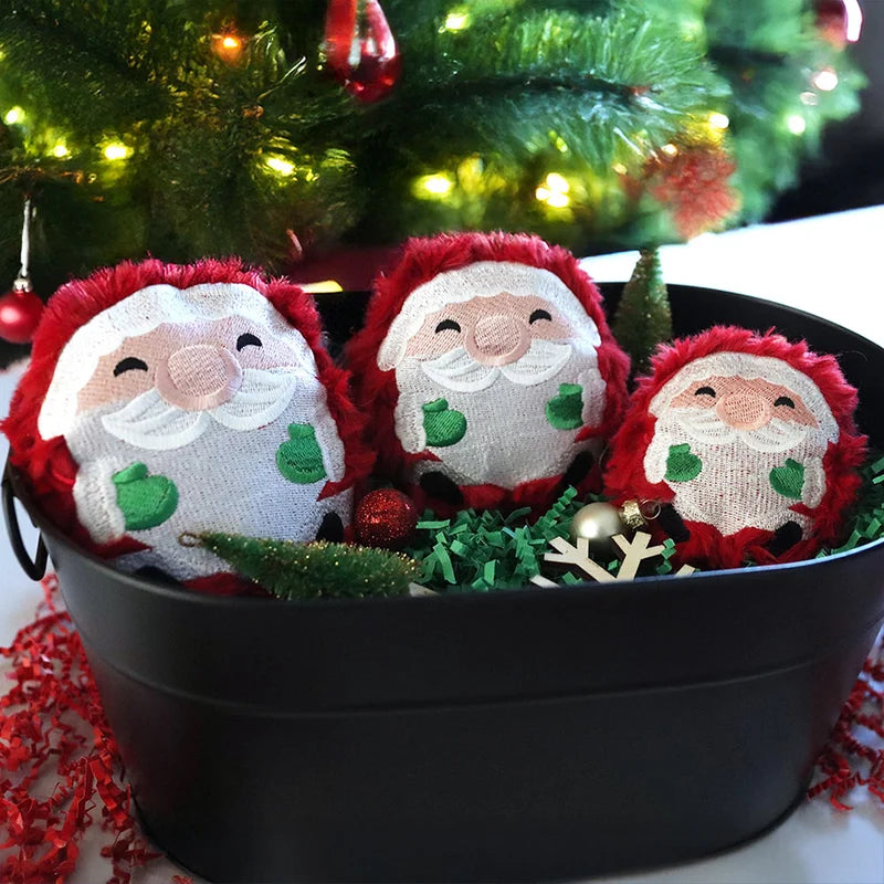 Three round embroidered Santa faces with red trim and green mittens sit in a black metal tub filled with green and red shredded paper, ornaments, and a small tree.