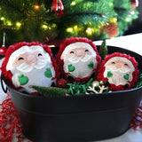 Three round embroidered Santa faces with red trim and green mittens sit in a black metal tub filled with green and red shredded paper, ornaments, and a small tree.