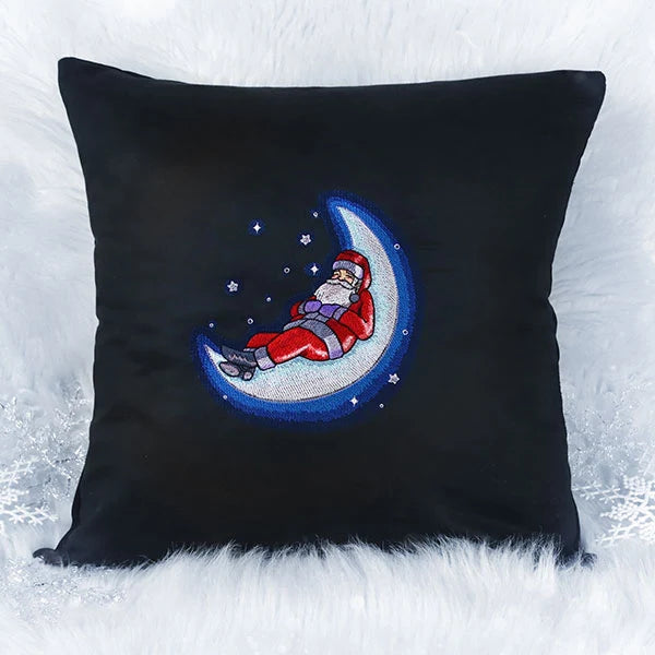 Black square pillow with embroidered design of Santa Claus in red suit lying on a glowing blue crescent moon surrounded by small stars.