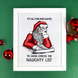Framed embroidery of Santa Claus in red and white with hand on face, next to text "IT'S ALL FUN AND GAMES 'TIL SANTA CHECKS THE NAUGHTY LIST" on white fabric.