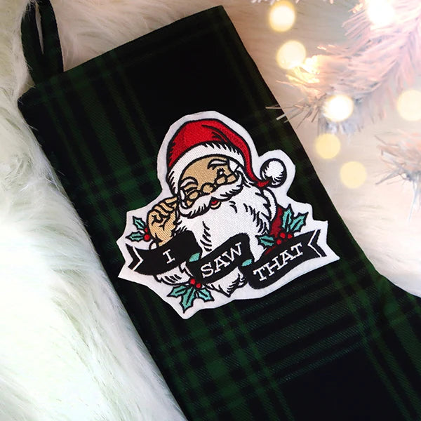 A dark green plaid Christmas stocking features an embroidered patch of Santa winking and holding glasses with the words I SAW THAT on a ribbon.