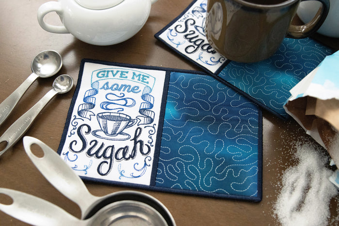 Machine embroidery in-the-hoop mug rug with text 'give me some sugah' next to kitchen elements.